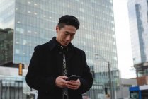 Vista frontal do jovem empresário asiático usando telefone celular na rua da cidade com prédio atrás dele — Fotografia de Stock