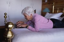 Seitenansicht einer aktiven Seniorin, die auf ihrem Bett liegt und zu Hause einen Laptop im Schlafzimmer benutzt — Stockfoto