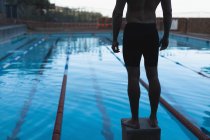 Section basse d'un nageur mâle debout sur le bloc de départ devant la piscine — Photo de stock