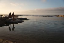 Vista lejana de la pareja afroamericana de pie y relajante cerca del mar en el atardecer. Están parados en la roca - foto de stock