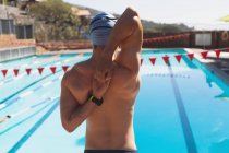 Vue arrière du jeune homme nageur caucasien étirant les bras tout en portant des maillots de bain et regarder à la piscine extérieure le jour ensoleillé — Photo de stock