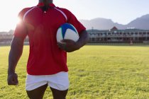 Sección media de un jugador de rugby masculino sosteniendo la pelota de rugby y de pie en el estadio en un día soleado - foto de stock