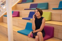 Vista frontal de uma bela empresária asiática sentada nas escadas e conversando em seu telefone celular no escritório — Fotografia de Stock