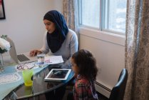 Вид збоку змішаної раси мати в хіджабі, використовуючи ноутбук, а дочка дивиться на цифровий планшет вдома. Вони сидять навколо столу у вітальні — стокове фото