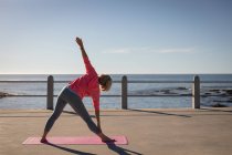 Visão traseira de uma mulher idosa ativa praticando exercícios de fitness em um tapete de fitness em um passeio ao sol em frente à praia — Fotografia de Stock