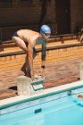 Frontansicht eines jungen kaukasischen männlichen Schwimmers, der am sonnigen Tag in Startposition im Schwimmbad auf dem Startblock steht — Stockfoto