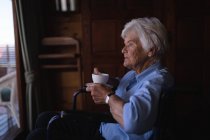 Vista lateral de uma mulher idosa ativa deficiente pensativa segurando um copo de café e sentada em uma cadeira de rodas no quarto em casa — Fotografia de Stock