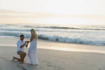 Vue latérale du bel homme caucasien mettant l'anneau dans le doigt de la femme à la plage — Photo de stock