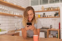 Вид спереду Жінка посміхається, використовуючи мобільний телефон вдома в кухонній кімнаті — стокове фото