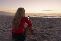 Вид сзади белокурой белокурой женщины с мобильного телефона на пляже. Она сидит на песке. — стоковое фото