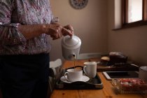 Sección media de una mujer mayor activa que vierte café en una taza en la mesa de comedor en la cocina en casa - foto de stock
