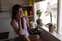 Frontansicht einer Frau mit gemischter Rasse, die mit dem Handy telefoniert, während sie zu Hause in der Küche einen Kaffee trinkt. sie lächelt — Stockfoto