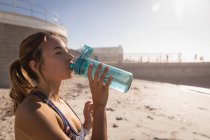 Vue latérale de la femme buvant de l'eau avec bouteille tout en se tenant à la plage par une journée ensoleillée — Photo de stock