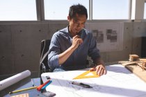 Front view of thoughtful Asian male architect working on blueprint at desk in a modern office. — Stock Photo