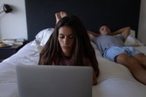 Vista frontal da bela mulher mestiça usando laptop enquanto o homem deitado na cama em casa — Fotografia de Stock