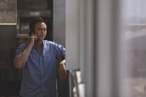 Vorderansicht eines männlichen asiatischen Architekten, der am Handy telefoniert, während er in einem modernen Büro am Fenster steht — Stockfoto