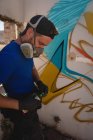 Vista lateral do jovem artista de grafite caucasiano de pé com pintura em spray na sala de beco — Fotografia de Stock