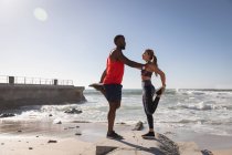 Vista laterale della giovane coppia multietnica che fa esercizio di stretching vicino alla spiaggia in una giornata di sole — Foto stock
