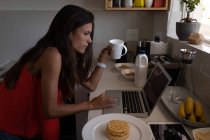 Vue latérale de belle femme mixte travaillant sur ordinateur portable tout en prenant un café dans la cuisine — Photo de stock