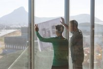 Rückansicht kaukasischer Architekten, die einen Bauplan gegen ein Fenster halten und im Büro darüber diskutieren — Stockfoto