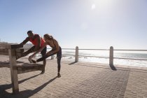 Vista frontal de una joven pareja multiétnica haciendo ejercicio de estiramiento en el banco cerca de la playa en un día soleado - foto de stock
