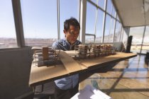 Vorderseite eines männlichen asiatischen Architekten, der ein architektonisches Gebäudemodell hält und am Schreibtisch in einem modernen Büro daran arbeitet — Stockfoto