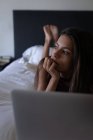 Front view of thoughtful mixed-race woman lying at bed while using laptop on bed at home. She is looking away — Stock Photo