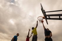 Visão de baixo ângulo de jogadores multi-étnicos jogando basquete na quadra de basquete contra um céu nublado — Fotografia de Stock