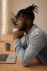 Side view of thoughtful African-American man with laptop sitting on chair at home. He is looking away — Stock Photo