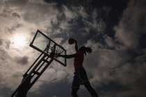 Visão de baixo ângulo do jogador de basquete afro-americano jogando basquete na quadra de basquete enquanto está pulando para marcar um afundanço contra o céu nublado escondendo sol — Fotografia de Stock