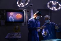 Вид спереду хірургів, що розмовляють один з одним під час операції в операційній кімнаті в лікарні проти вогнів і цифрового екрану — стокове фото