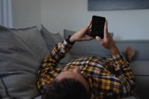 Vista trasera del joven hombre caucásico utilizando el teléfono móvil mientras está acostado en el sofá en casa - foto de stock