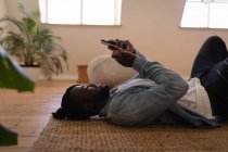 Вид сбоку на красивого афроамериканца, который пользуется мобильным телефоном, лежа дома на полу — стоковое фото