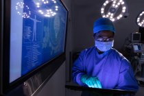 Вид спереду чоловічого хірурга з використанням цифрового планшета в операційній кімнаті в лікарні з великим цифровим екраном позаду нього — стокове фото