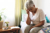 Vorderansicht einer traurigen Seniorin, die im Altersheim auf dem Bett sitzt. Frau sieht frustriert mit Hand auf der Stirn aus. — Stockfoto