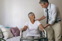 Front view of male Caucasian doctor helping senior mixed race female patient to walk with walker at retirement home — Stock Photo