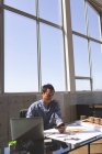 Vista frontal del reflexivo arquitecto asiático sentado en el escritorio mientras usa el teléfono móvil en una oficina moderna. Regla de triángulo naranja, brújula de geometría, lápiz, computadora portátil y modelo arquitectónico se muestran en el escritorio en la oficina moderna . - foto de stock
