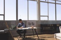 Vista frontal del trabajador arquitecto asiático sentado en el escritorio y trabajando en el ordenador portátil en una oficina moderna contra el cielo azul en el fondo - foto de stock