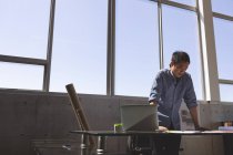 Вид азиатского архитектора-мужчины, стоящего за рабочим столом и выглядящего синим в современном офисе — стоковое фото