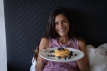 Portrait of beautiful mixed-race woman holding plate of breakfast while leaning on bed at home. She is looking and smiling at camera — Stock Photo