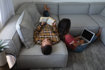 Visão traseira da mulher mestiça usando laptop enquanto homem caucasiano livro de leitura em casa — Fotografia de Stock