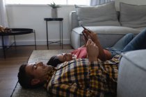 Вид сбоку на молодую многоэтническую пару, которая пользуется мобильным телефоном, отдыхая дома на полу — стоковое фото