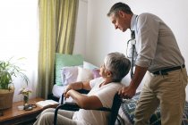 Vue latérale du médecin masculin caucasien interagissant avec une patiente âgée de race mixte à la maison de retraite — Photo de stock