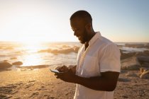 Vista lateral do jovem afro-americano usando telefone celular na praia ao pôr do sol. Ele está sorrindo. — Fotografia de Stock