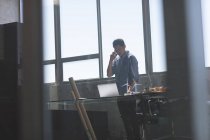 Вигляд спереду азіатських чоловічого архітектора стояти і говорити на мобільному телефоні під час роботи над столом у сучасні офісні — стокове фото