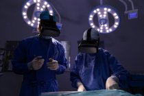 Visão frontal de cirurgiões que utilizam fones de ouvido de realidade virtual durante a cirurgia no centro cirúrgico do hospital com pontos acima deles — Fotografia de Stock