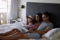 Vue latérale du couple romantique multi-ethnique à l'aide d'un ordinateur portable tout en étant couché sur le lit à la maison. Ils sourient. — Photo de stock