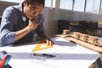 Front view of thoughtful Asian male architect working on blueprint while touching chin with hand at desk in a modern office — Stock Photo