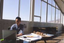 Front view of a hardworking Asian male architect using laptop while working on blueprint with orange triangle ruler, geometry compass and pencils at desk in a modern office against blue sky in background — Stock Photo