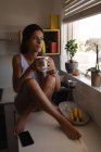 Front view of beautiful mixed-race woman drinking coffee while sitting in kitchen at home. She is looking away — Stock Photo